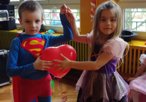 Chłopiec przebrany za Spidermana i dzieczynka-czarownica trzymają między sobą balon w kształcie serca.